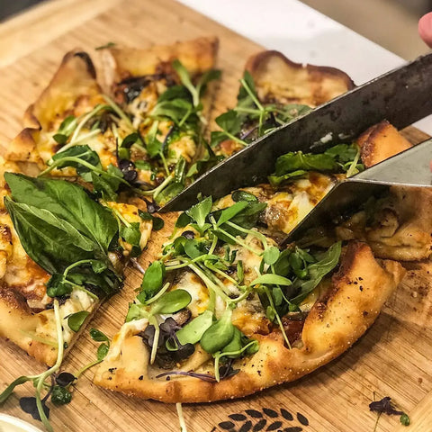 A homemade pizza topped with cheese, greens, basil, and Gustus Vitae Greek Seasoning Gift Set’s artisanal blends is being sliced with a large knife on a wooden cutting board.