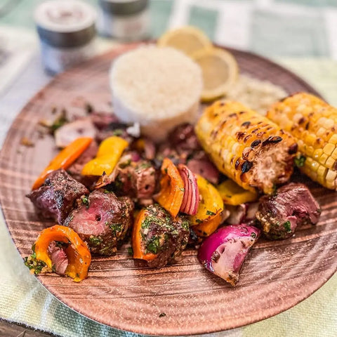 A wooden plate holds grilled steak, vegetable skewers, corn on the cob, white rice, and lemon slices. In the background, jars of Gustus Vitae’s Greek Seasoning Gift Set - Tastes of Greece artisanal spice blends are visible.