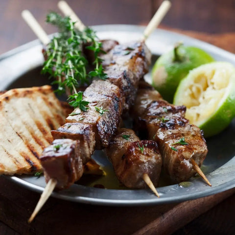 Three grilled meat skewers, seasoned with Gustus Vitaes Middle Eastern Seasoning Gift Set, garnished with fresh herbs and served with grilled bread and lime on a metal plate atop a rustic table.