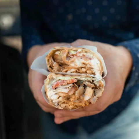 A person in a dark blue dotted shirt holds a halved sandwich filled with sliced chicken, tomatoes, and creamy sauce enhanced by the Gustus Vitae Middle Eastern Seasoning Gift Set—artisan spice blends inspired by Tastes of the Middle East.
