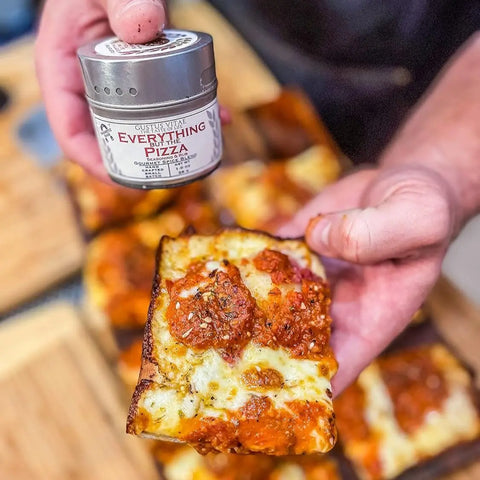 A person holds a cheesy pizza slice in one hand and a small container from the Gustus Vitae Everything but the Pizza Night Collection | Set of 4 in the other, with more pizza slices visible in the background.