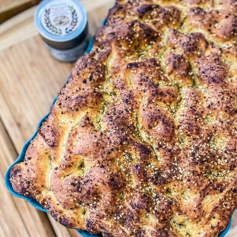 A golden-brown, herb-topped focaccia in a blue dish rests on wood, with the Gustus Vitae Everything but the Pizza Night Collection | Set of 4 from our artisanal spice lineup nearby in the background.