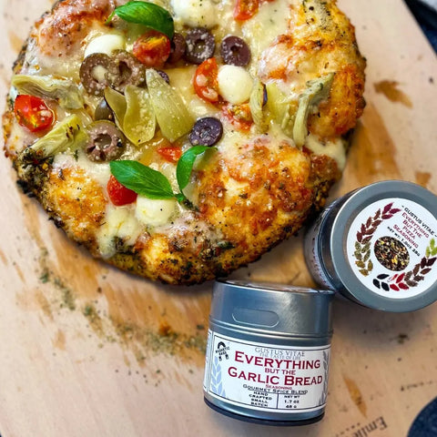 A round, cheesy veggie pizza with olives, tomatoes, artichokes, and basil sits on a wooden surface beside Gustus Vitaes Everything but the Pizza Night Collection | Set of 4 artisanal seasonings.