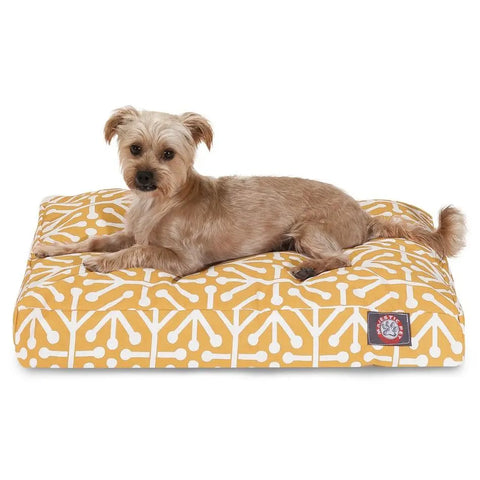 A small, tan dog rests on the Majestic Pet Aruba Rectangle Dog Bed, which features a yellow and white geometric pattern, a visible brand patch, and a machine washable cover. The dog gazes at the camera, highlighting the beds comfort and style.