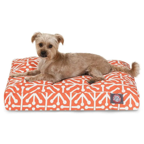 A small, tan dog lies on the Majestic Pet Aruba Rectangle Dog Bed with a white geometric pattern; its machine washable cover and premium polyester fiber fill offer comfort as the dog gazes at the camera.