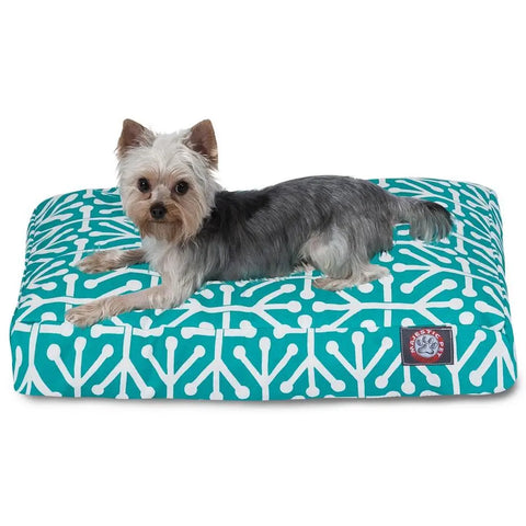 A small Yorkshire Terrier lies on the Majestic Pet Aruba Rectangle Dog Bed, featuring a teal and white pattern, a red and white logo patch in one corner, and a machine-washable cover for easy cleaning.