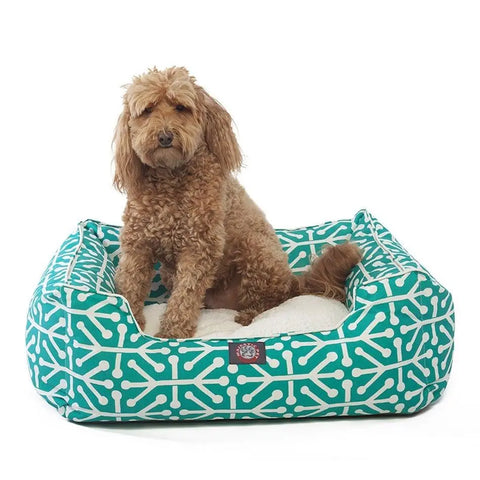 A curly-haired brown dog relaxes on the Majestic Pet Aruba Contemporary Lounge Bed—a rectangular teal bed with a white geometric pattern and a soft, machine-washable white cushion.