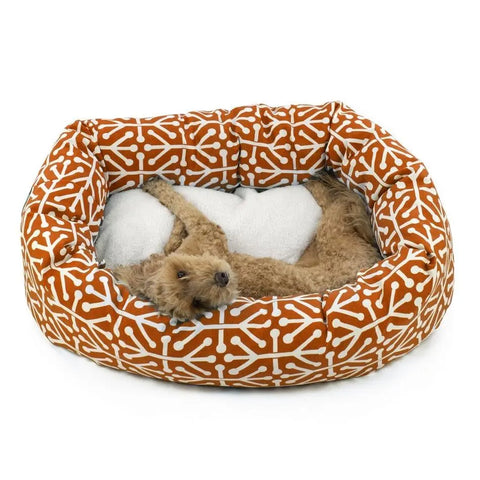 A brown curly-haired dog lies on its back with front paws outstretched in the Majestic Pet Aruba Sherpa Bagel Dog Bed, which features an orange and white pattern and orthopedic spine support.