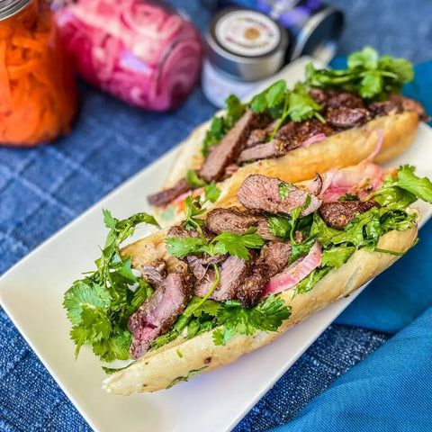 Two banh mi sandwiches with grilled steak seasoned using Gustus Vitae’s Asian BBQ Seasonings Collection - 3 Pack, fresh cilantro, and pickled veggies are served on a white plate, with jars of condiments in the background on a blue surface.