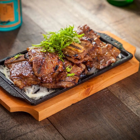 Grilled marinated beef short ribs served on a sizzling cast iron plate, topped with sliced green onions and Gustus Vitae’s Asian BBQ Seasonings Collection - 3 Pack, presented on a wooden tray on a rustic table.