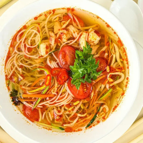 A steaming bowl of noodle soup, topped with tomatoes, chili peppers, veggies, and herbs in a clear broth seasoned with Gustus Vitaes Asian Seasonings Gift Set - Tastes of Asia artisanal spice blends. Parsley garnish; spoon on the side.
