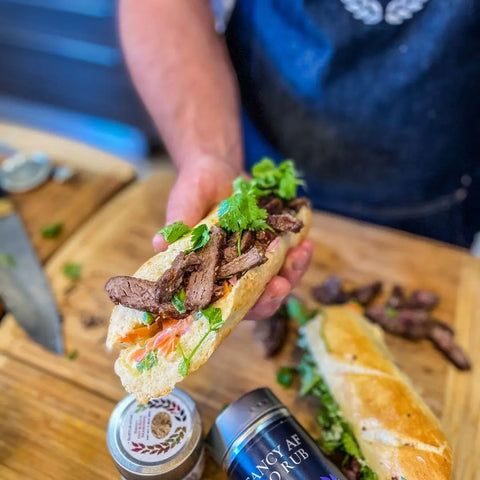 A person holds a fresh baguette sandwich with sliced steak, greens, and veggies over a wooden board, with the Gustus Vitae Asian Seasonings Gift Set—Tastes of Asia artisanal spice blends six pack and another sandwich nearby.
