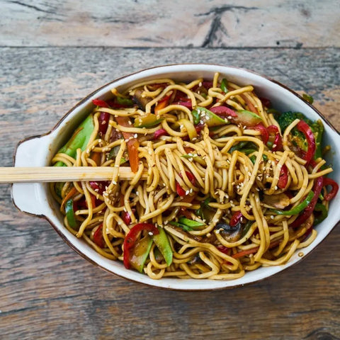 A white oval dish of stir-fried noodles and veggies with sesame seeds, seasoned with Gustus Vitae’s Asian Seasonings Gift Set—Tastes of Asia Six Pack. Chopsticks rest on top, and the dish sits on a rustic wooden surface.