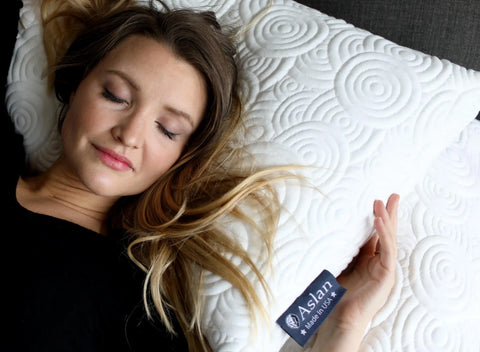 A woman with long blonde hair sleeps peacefully on the Aslan Adjustable Pillow by Aslan Mattress, featuring a cool-to-the-touch, white textured cover with circular patterns. She wears a black top, and the pillow bears the Aslan Mattress label.