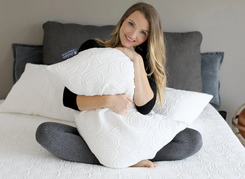 A woman sits cross-legged on a bed, smiling and hugging the Aslan Adjustable Pillow by Aslan Mattress, which features a geometric swirl pattern and a cool to the touch cover. The bed has white bedding and gray pillows in the background.
