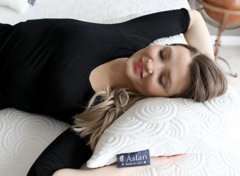 A woman in a black shirt relaxes, eyes closed and smiling, on a white patterned memory foam mattress and the Aslan Adjustable Pillow by Aslan Mattress.