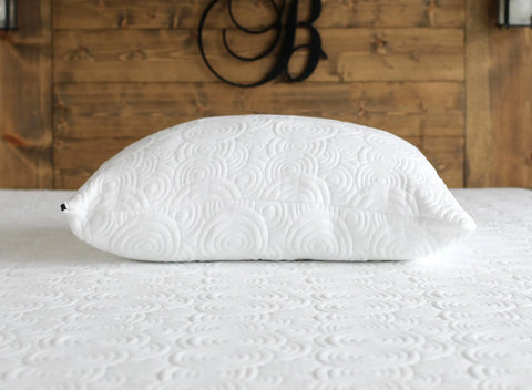 The Aslan Adjustable Pillow by Aslan Mattress, featuring a raised circular pattern, rests on a matching textured mattress with a wooden headboard and a decorative letter B in the background.