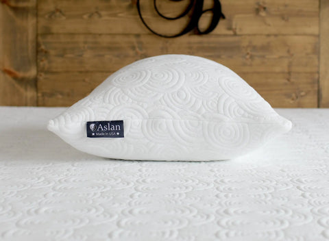 The Aslan Adjustable Pillow by Aslan Mattress, with a cool-to-the-touch white cover and textured circular pattern, rests atop a matching bedspread, set against a warm wooden wall.