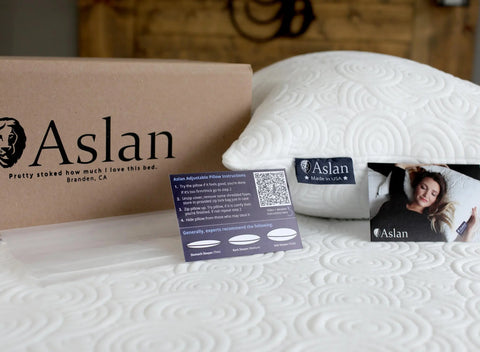A white Aslan Adjustable Pillow by Aslan Mattress with instruction cards sits on a textured white bed. A brown Aslan Mattress box is nearby, and one card features a woman sleeping on the pillow’s cool-to-the-touch cover.