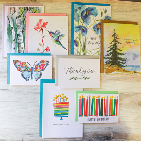The JuLu Cards Assorted Card Bundle includes eight colorful greeting cards with watercolor designs—flowers, a tree, butterfly, birthday cake, candles, and messages like Thank you and With Deepest Sympathy—arranged on light wood.