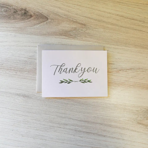 A white JuLu Cards thank you card with cursive script and a small green leaf, from the Assorted Card Bundle, sits atop its matching envelope on a light wooden surface—ideal for sympathy notes or your greeting card collection.