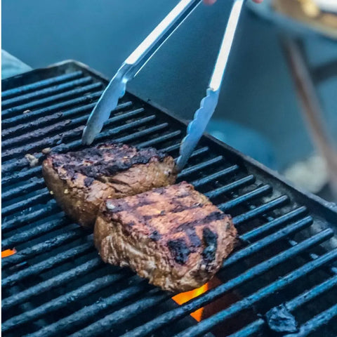 A set of steaks sizzles over an open flame, with metal tongs gripping one piece. Distinct grill marks and BBQ Pit Master | Set of 4 seasoning from vendor-unknown enhance the look, while seasoned grates hint at past cookouts.