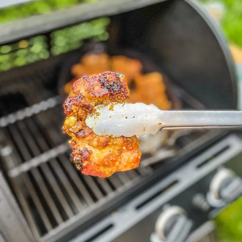 Using the vendor-unknown BBQ Pit Master | Set of 4, a piece of grilled, seasoned chicken is held with metal tongs above an open barbecue grill, while more chicken cooks and sizzles in the background.