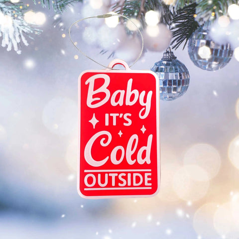 The Chatelet Manufacturing Baby Its Cold Outside Christmas Ornament—decorative and made in the USA—hangs on a snowy tree branch, displaying the festive phrase in white amid silver baubles and glowing holiday lights.