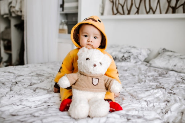 Child in a yellow raincoat holding a white teddy bear with a brown sweater on a bed.