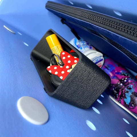 A close-up of a blue bag’s interior shows the Chatelet Manufacturing BOGLETS Organizer Charm Accessory for Bogg Bags, holding a keychain with a red and white polka dot bow, lip balm, a bottle, and other small patterned items.