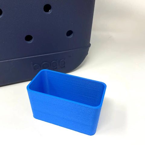 A small, rectangular blue BOGLETS organizer charm accessory by Chatelet Manufacturing sits in front of a navy Bogg Bag with round holes and bogg embossed on it, against a white background. Perfect for keeping keys or personal items handy.