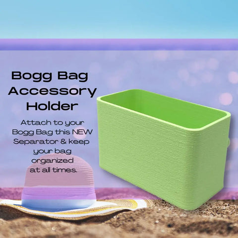 A green BOGLETS Organizer Charm Accessory by Chatelet Manufacturing is shown on a beach with a pastel-striped hat and sunglasses. It keeps keys or personal items handy with your Bogg Bag tote, helping you stay organized.
