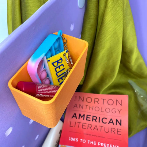 A yellow BOGLETS Organizer Charm Accessory by Chatelet Manufacturing holds lip balm, a pop-it keychain, and a BELIEVE keychain—ideal for keeping keys or personal items handy with your tote bag or Bogg Bag. Nearby: a green cloth and The Norton Anthology.