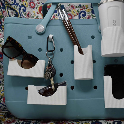 A Chatelet Manufacturing BAGLETS Ultimate Bundle in white is displayed on a teal perforated tote, featuring a phone case holder, key & wallet holder, carabiner key holder, bottle holder, and sunglasses holder with accessories against a floral background.
