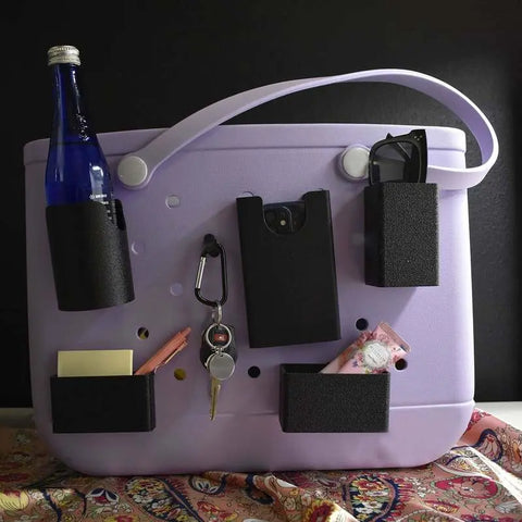 A lavender tote stands upright with Chatelet Manufacturing’s BAGLETS Ultimate Bundle—black phone case holder, key & wallet holder, carabiner key holder, bottle and sunglasses holders—attached to carry essentials. The bag rests on a colorful patterned cloth.