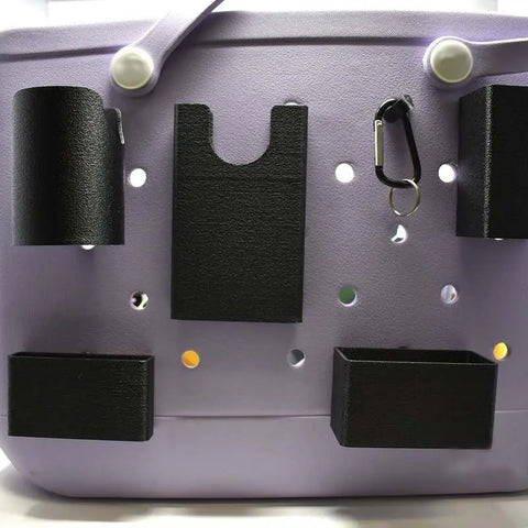 A light purple tote bag with Chatelet Manufacturings BAGLETS Ultimate Bundle—black phone case holder, key & wallet holder, carabiner key holder, bottle holder accessory, and sunglasses holder—attached to its front with multiple circular holes.