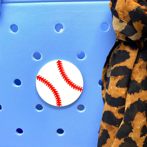 The Chatelet Manufacturing BOGLETS Sport Charm, a round pin with red baseball stitching, is attached to a light blue rubber beach bag near leopard print fabric—perfect for customizing your tote or Bogg Bag.