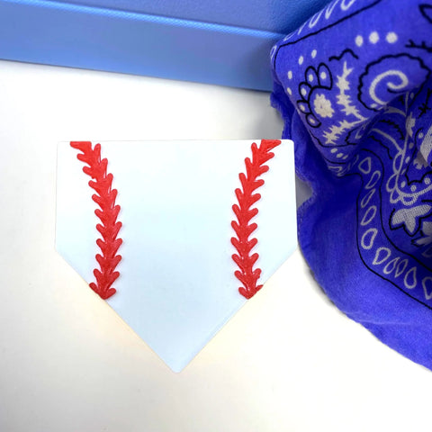 A white home plate-shaped BOGLET Sport Charm with red baseball stitching by Chatelet Manufacturing sits near a light blue box and a blue paisley bandana—perfect for adding sporty flair to your rubber beach or tote bags.