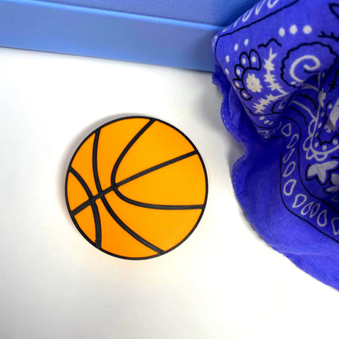 A flat, round basketball-themed charm from Chatelet Manufacturing’s BOGLETS—Sport Charms for Rubber Beach Bags—rests on a white surface by a blue item and paisley fabric, ideal for personalizing your tote or Bogg Bag accessories.