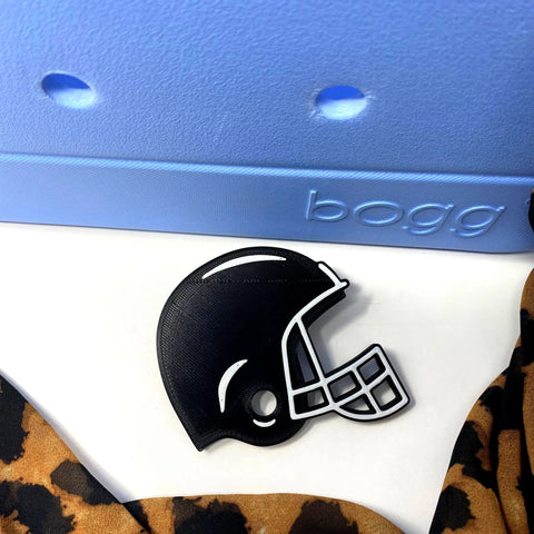 A black football helmet-shaped BOGLET Sport Charm by Chatelet Manufacturing sits on a white surface near a blue bogg bag and leopard print fabric—perfect for customizing rubber beach bags with unique accessories.