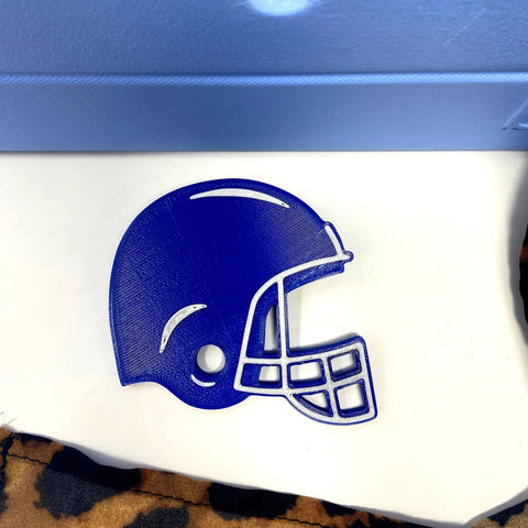 A blue football helmet BOGLET Sport Charm by Chatelet Manufacturing, designed for rubber beach bags and tote bags, sits on a white surface near a light blue wall and brown patterned fabric.