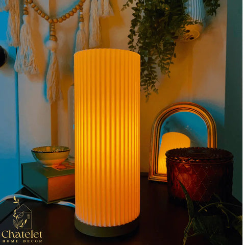 The Beam Lamp from Chatelet Manufacturing’s Premium Collection casts a warm, color-changing glow on a dark table beside a round mirror, decorative box, books, beads, and green plants. The Chatelet Manufacturing logo appears in the corner.