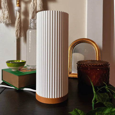 The Beam Lamp by Chatelet Manufacturing, a white ribbed color-changing light with a tan base, elevates premium decor on a dark tabletop alongside a green bowl on a book, glass bottle, gold mirror, decorative box, and green leaves.