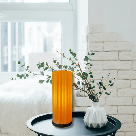 The Beam Lamp by Chatelet Manufacturing, featuring premium ambience lighting, sits alongside a white vase with green branches on a round black tray table in a cozy, bright room with white brick walls and a window.