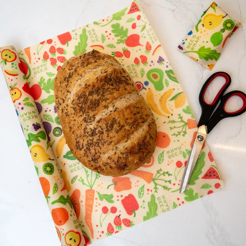 A loaf of bread rests on Bees Wrap Roll, a colorful, patterned beeswax food wrap—an eco-friendly plastic wrap alternative for home bakers. Scissors and a folded piece of the same wrap sit nearby on the white countertop.