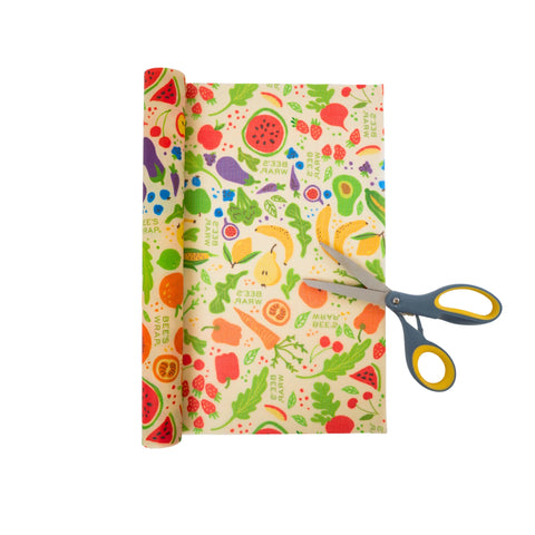A Bees Wrap Roll featuring a fruit and vegetable design is partially unrolled on a white background—a sustainable alternative to plastic wrap. Yellow and gray scissors are placed beside the roll, ready for cutting.