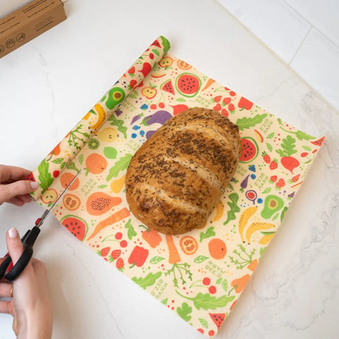 A person uses scissors to cut a Bees Wrap Roll, featuring a colorful fruit and veggie pattern, to fit around a loaf of bread. This reusable wrap by Bees Wrap is a perfect plastic wrap alternative for home bakers.