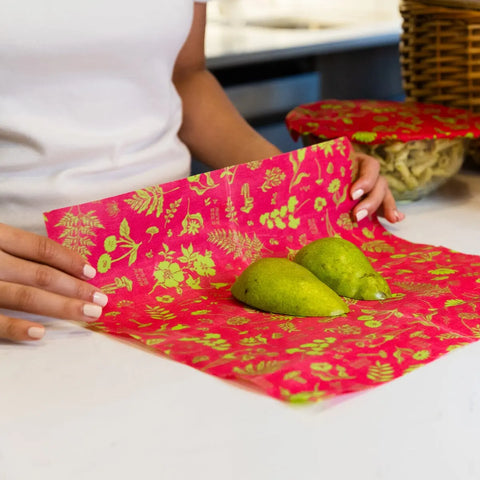 On a kitchen counter, someone wraps two green pears in Bees Wrap Roll, a red and yellow patterned reusable food wrap by Bees Wrap—a perfect plastic wrap alternative. In the background are a covered bowl and wicker basket.