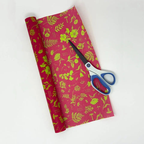 A pair of scissors rests on a Bees Wrap Roll—red wrapping paper with yellow floral and botanical patterns—offering a customizable format ideal for home bakers, set against a white background.