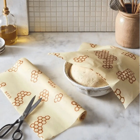 A Bees Wrap Roll, scissors, and a bowl covered with honeycomb-patterned Bees Wrap—a plastic wrap alternative—sit on a marble countertop beside a jar of utensils and a bottle of oil.